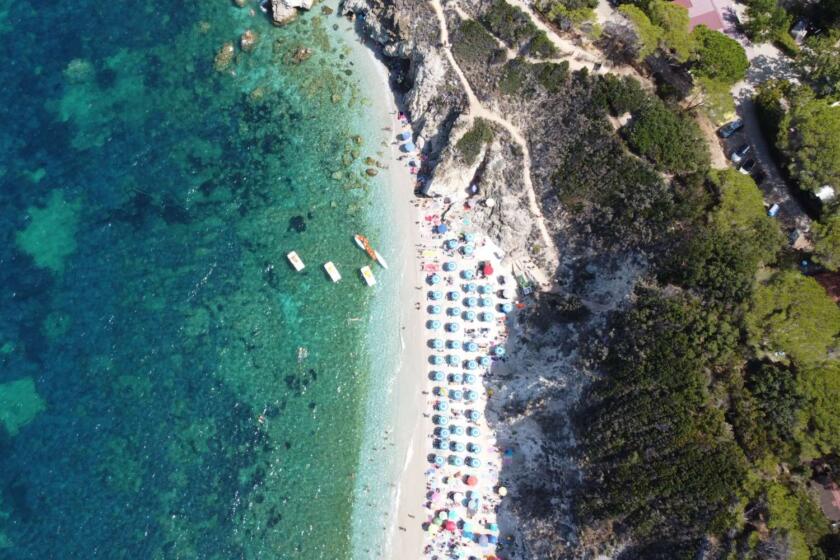 Spiaggia dell’Arcipelago Toscano vista dall’alto con mare cristallino, ombrelloni colorati e costa rocciosa mediterranea

